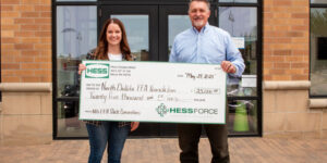 Laiken DeMorrett and Brent Lohnes stand smiling in front of the Hess office in Minot, North Dakota, holding a large ceremonial check for $25,000 made out to the North Dakota FFA Foundation, dated May 29, 2025.